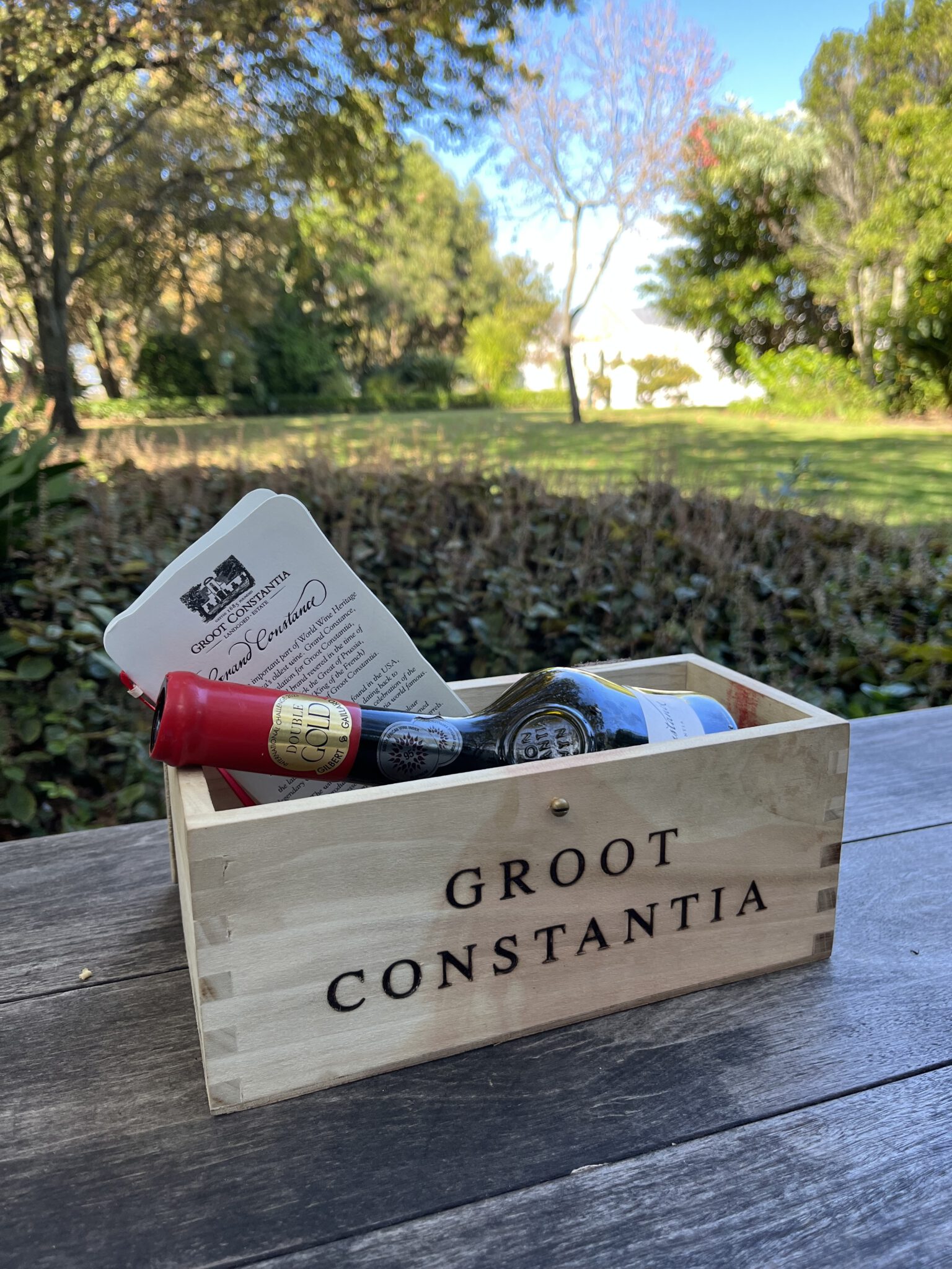Holzkiste mit Groot Constantia Aufschrift und Grand Constance Weinflasche auf einem Holztisch im Garten